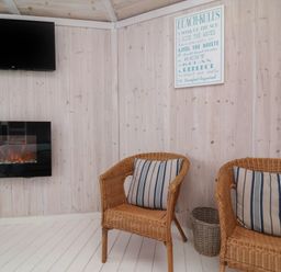A room with two wicker chairs with striped cushions a wall-mounted electric fireplace a wall-mounted tv and a beach rules sign at Shipload Bay near Hartland