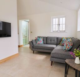 A living room with a grey sectional sofa with colorful pillows a small wooden side table with flowers and a wall-mounted flat-screen TV at Upper Cottage in Rocombe near Uplyme