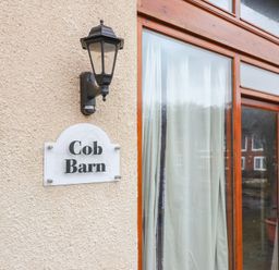 A wall with a light fixture and a sign that says Cob Barn next to a wooden framed glass door with curtains at Cob Barn in Aylesbeare