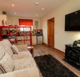 A living room with a sofa, black rug, electric stove heater, wall-mounted TV, red shelving unit, and small kitchen at Stable Cottage in Cottonshopeburnfoot near Otterburn