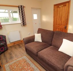 A living room with a brown sofa with white cushions a patterned armchair wooden door and window with curtains at Fir Tree Cottage in Cottonshopeburnfoot near Otterburn