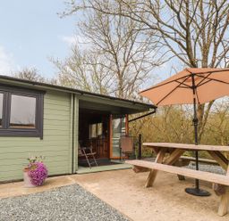 An outdoor patio area with a wooden picnic table and umbrella next to a green wooden cabin at Mill Cabin Denant in Haverfordwest