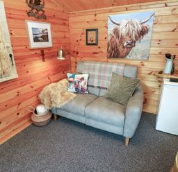 A small room with wooden walls featuring a grey two-seater sofa with cushions and a throw a basket under the sofa a mini fridge with kitchen utensils and a microwave on the counter at Mill Cabin Denant in Haverfordwest