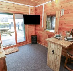 A wooden cabin interior with a small dining table set for two a wall mounted television glass door and a picnic table outside at Mill Cabin Denant in Haverfordwest