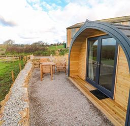 A wooden pod with glass double doors and outdoor wooden furniture on gravel near a stone wall at Audrey in Penzance near Newlyn