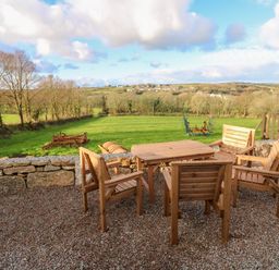 An outdoor seating area with wooden table and chairs overlooking a green field with trees and farm equipment at Lloyd in Penzance near Newlyn