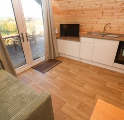 A small kitchen area with white cabinets a sink a black oven and a television next to glass doors with curtains and a green sofa at Lloyd in Penzance near Newlyn