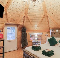A wooden bedroom with a bed, green pillows and towels, a bedside table with a lamp, a door, a wall-mounted TV, and a hanging robe at Hexagon Hut - Dec Update in Dungiven Londonderry