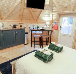 A small wooden room with a bed, green towels, a kitchenette with microwave and kettle, a high table with stools, and a door with a window at Hexagon Hut - Dec Update in Dungiven Londonderry