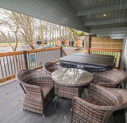 A covered outdoor deck with a round glass table and wicker chairs next to a hot tub overlooking a parking area and trees at Mill View in Dungiven Londonderry