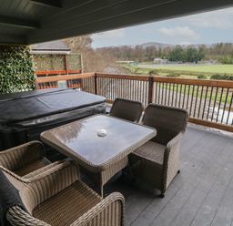An outdoor covered patio with a hot tub a glass table and four wicker chairs overlooking a grassy field and trees at The Roe in Dungiven Londonderry