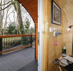 A wooden balcony with railing overlooking trees and a grassy area next to an interior space with wooden walls a small table with a bottle and remote and a floor lamp Hazel Dungiven Londonderry