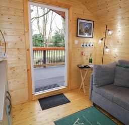 Interior view of a wooden cabin with a gray sofa a side table with a bottle and a lamp and an open door leading to a deck at Hazel in Dungiven Londonderry