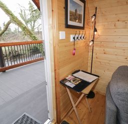 A wooden interior corner with a gray couch a small table with a binder and remote control a floor lamp and an open door leading to a deck over a river at Juniper in Dungiven Londonderry
