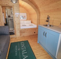 A small wooden cabin interior with a bed, shower, grey sofa, blue kitchenette, and a green floor mat at Juniper in Dungiven Londonderry