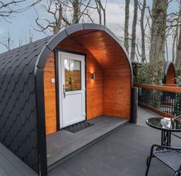 An outdoor pod with a white door and wooden interior on a deck with two black chairs and a round glass table at Birch in Dungiven Londonderry