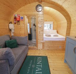 An interior of a wooden cabin with a gray sofa green pillows a bed with folded towels a small bathroom and a kitchenette at Kintail Resort Dungiven Londonderry