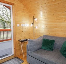 A room with wooden walls featuring a grey sofa with green cushions a small side table with reading materials and a floor lamp next to an open door leading to an outdoor area at Yew in Dungiven Londonderry