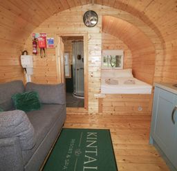 A wooden cabin interior with a gray sofa green cushions kitchenette bed with towels and a shower at Kintail Resort Spa
