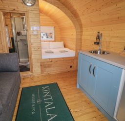 A wooden cabin interior with a gray sofa a bed shower area a blue kitchenette and a green rug at Kintala Resort and Spa