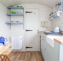 A small kitchen with wooden floors wooden shelves with dishes and glasses a wooden table with a blue teapot and mugs a door in the center and a kitchen sink on the right at Teg in Prospidnick near Porthleven