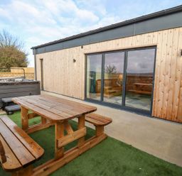 An outdoor area with a wooden picnic table and benches next to a hot tub by a building with wooden paneling and sliding glass doors at Bears Court Barn 4 in Little Rissington near Bourton-On-The-Water