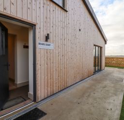 Exterior view of a wooden building with an open black door and a sign that reads Bears Court 4 at Little Rissington near Bourton-On-The-Water