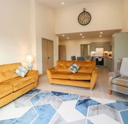 A living room with two orange sofas a plaid armchair a large clock on the wall and a kitchen in the background at Bears Court Barn 4 Little Rissington near Bourton-On-The-Water
