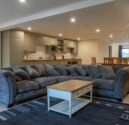 An open plan room with a grey sectional sofa coffee table kitchen cabinets and a dining table with chairs at Bears Court Barn 8 Little Rissington near Bourton-On-The-Water