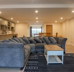 An open plan living room with a grey sectional sofa a white coffee table a dark patterned rug a dining table with wooden chairs and a kitchen with grey cabinets at Bears Court Barn 8 in Little Rissington near Bourton-On-The-Water
