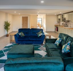 An open-plan living room with blue and green sofas a patterned rug wooden side tables a kitchen area with cabinets and appliances and a dining area in the background at Bears Court Barn 5 in Little Rissington near Bourton-On-The-Water