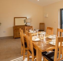 A dining room with a wooden table set for six with bowls and glasses a wooden sideboard with a lamp a plant in the corner and a glass door letting in natural light at Bears Court Barn 5 Little Rissington near Bourton-On-The-Water
