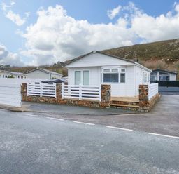 A white single-story house with a stone and white fence on a corner lot with a paved driveway and hills in the background at Un Diwrnod (One Day) The Warren T22 The Warren near Abersoch