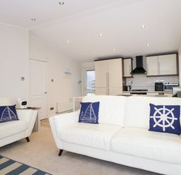 A living room with white sofas and blue nautical pillows facing a kitchen with cream cabinets at Un Diwrnod The Warren T22 The Warren near Abersoch