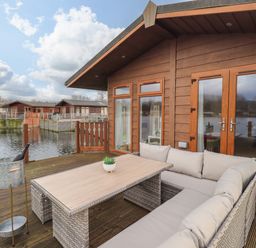 An outdoor deck with a wicker sectional sofa and table next to a wooden cabin by the water at Otters Reach near Carnforth