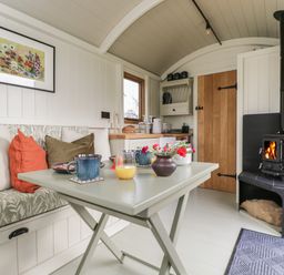 A small living space with a foldable table set with cups and flowers a built in bench with cushions a wood burning stove and a kitchen area at Blackthorn in South Leigh near Witney