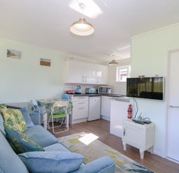 A living area with a blue couch green patterned cushions a small dining table with four chairs a white kitchen with appliances a wall-mounted TV and a small cabinet at Number 19 in Cromer