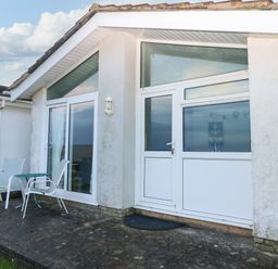 An exterior view of a building with white walls glass doors windows and outdoor chairs with a table on a concrete patio at Chalet 27 in Port Eynon