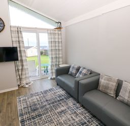 A living room with two gray sofas and patterned cushions a wall clock a mounted TV and glass doors leading outside at Chalet 27 in Port Eynon