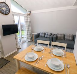 A dining table set with plates bowls glasses and cutlery in a room with two sofas a wall clock and a mounted television at Chalet 27 in Port Eynon