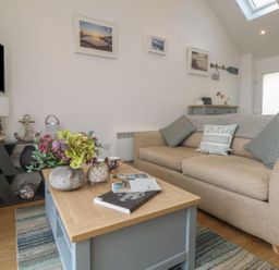 A living room with a beige sofa and cushions a wooden coffee table with flowers a TV on a stand and framed pictures on the wall at The Loft in Porthleven