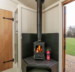 A wood burning stove with a fire and a fire extinguisher inside a small room with a wooden door and open door to a garden at Hazel in South Leigh near Witney