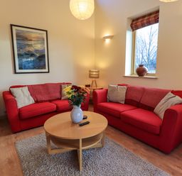 A living room with red sofas a round wooden table a vase with flowers and a window at Larch Lodge in Skelton near Penrith