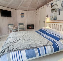 A bedroom with a bed with striped bedding two chairs a wall-mounted television and an electric fireplace at Barley Bay near Hartland