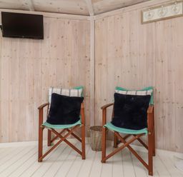 Two wooden chairs with cushions against a wooden paneled wall with a wall mounted tv and a white tower fan at Spekes Mill near Hartland