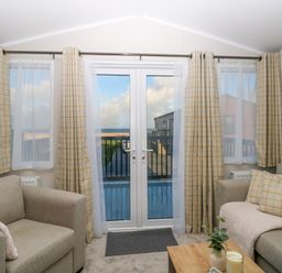 A living room with two sofas a small wooden table and glass doors with curtains at The Sea Lodge in Bacton near Walcott