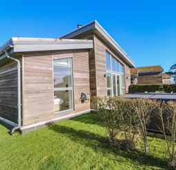 An exterior view of a wooden house with large windows and a covered hot tub on a grassy yard at Una Lodge 77 in Carbis Bay