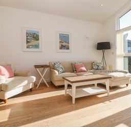 A living room with beige sofas a wooden coffee table paintings on the wall and large windows at Una Lodge 77 in Carbis Bay