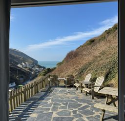 A stone patio with wooden chairs and tables overlooking a hillside and a coastal village at Little Ruffo Trebarwith near Tintagel