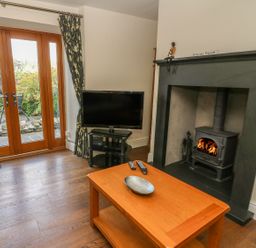 A living room with a wood stove fireplace television on a stand wooden coffee table with remotes and glass doors to a garden at Woodbine Cottage in Ambleside
